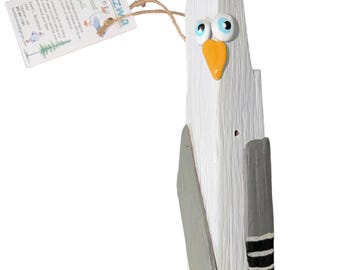 Könnte beinhalten: Eine handgefertigte Möwenfigur aus weiß bemaltem Treibholz mit einem leuchtend orangefarbenen Schnabel und blau-weißen Augen. Sie hat grau bemalte Flügel mit schwarz-weiß gestreiften Spitzen. Ein Papieranhänger mit der Aufschrift Kemizmz designs ist befestigt.