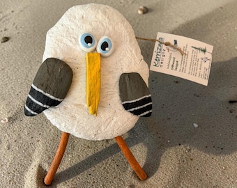 Puede incluir: Una figura decorativa de gaviota hecha con un cuerpo de piedra blanca texturizada, un pico de madera amarillo brillante y dos ojos con borde azul. Presenta alas de color gris oscuro con rayas blancas y patas de palito de color naranja.