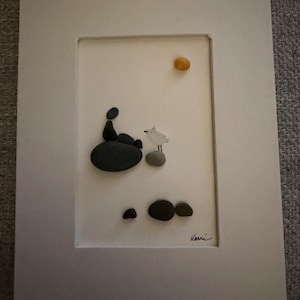 May include: Framed pebble art depicting a person and a bird on a rock, with a yellow sun in the sky. The artwork is signed "Kerri" in the bottom right corner. The frame is white.