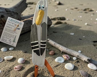 Könnte beinhalten: Eine Treibholz-Möwen-Skulptur mit weißem Körper, grauen Flügeln und orangefarbenen Beinen steht an einem Sandstrand. Die Möwe hat einen gelben Schnabel und blaue Augen. Ein Etikett lautet "Kerrizwa designz driftwood seagull."
