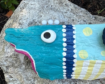 Könnte beinhalten: Handbemalte Holzfischskulptur in Türkis-, Blau- und Gelbtönen. Der Fisch hat ein großes weißes Auge mit schwarzer Pupille, weiße Punkte und gelbe Streifen. Die Skulptur liegt auf einem grauen Felsen.
