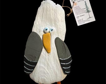 Könnte beinhalten: Eine handgefertigte Möwenfigur aus bemalten Steinen und Treibholz mit weißem Körper, grauen Flügeln mit schwarzen Streifen, gelbem Schnabel und zwei kleinen Wackelaugen. Ein weißes Etikett mit der Aufschrift Kerrizma designz hängt an einer braunen Schnur.