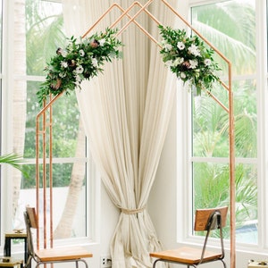 May include: A copper wedding arch with white and green floral arrangements on each side. Two wooden chairs with metal frames are placed in front of the arch.