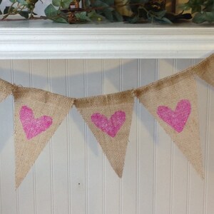 May include: A burlap banner with pink glitter hearts. The banner is strung together with twine and is hanging on a white wall.