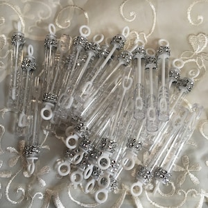 May include: A group of clear plastic bubble wands with silver glitter and white handles. The wands are arranged in a pile on a white lace background.