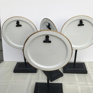 May include: Four white round plates with gold trim and black clips are mounted on black stands. The stands have a square base.