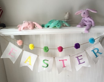 May include: A white banner with the word "EASTER" spelled out in colorful letters hangs from a string with colorful pom-poms. Three plush bunny rabbits in pink, green, and purple are sitting on a white shelf.