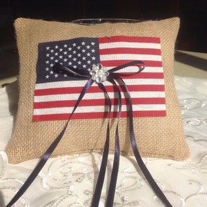 May include: A burlap ring pillow with an American flag design and a black satin ribbon with a rhinestone embellishment.