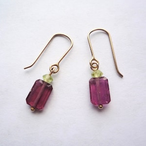 May include: A pair of gold-colored earrings. Each earring features a rectangular, deep pink gemstone with a small, light green gemstone above it. The earrings have a simple hook design.