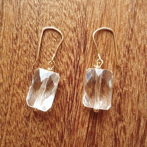 May include: A pair of gold-colored earrings with clear, faceted rectangular crystal pendants. The earrings have a simple hook design and are displayed against a warm, wooden background.