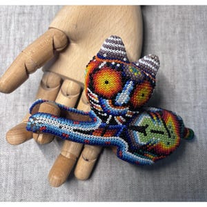 May include: A colorful beaded cat figurine with a long tail. The cat is adorned with orange, yellow, blue, and green beads. The figurine is held in a wooden hand, showcasing the intricate beadwork and design.
