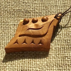 Pode incluir: Um pingente de madeira com um rosto esculpido e um cordão de couro marrom. O rosto tem uma boca esculpida, olhos e uma fileira de triângulos na parte inferior.