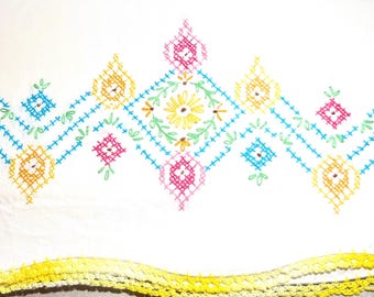 May include: White fabric with colorful cross-stitch embroidery. The design features a central floral motif with yellow, pink, and green accents, surrounded by geometric shapes in blue, yellow, and pink. A yellow crocheted trim is at the bottom.