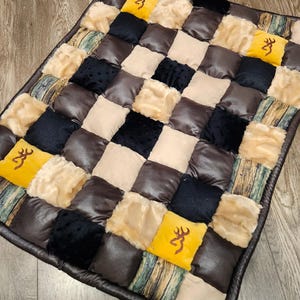 May include: A rectangular patchwork quilt featuring a grid of alternating squares in brown faux leather, black textured fabric, beige faux fur, and wood-grain patterned fabric. Several bright yellow squares feature a brown deer antler logo.