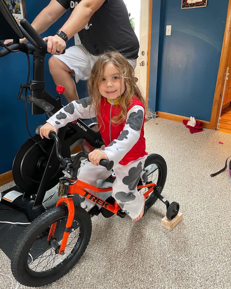 May include: A young girl wearing a red and white dalmatian print shirt is riding a bright orange Trek bicycle with training wheels. The bicycle has black tires and a black seat. The girl is smiling and looking at the camera. The bicycle is in front of a stationary exercise bike.