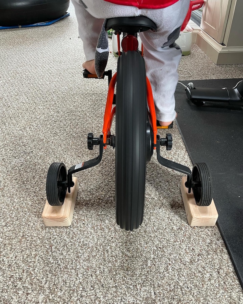 May include: A red and black children's bicycle with training wheels on wooden blocks. The bicycle has a black seat and black handlebars.