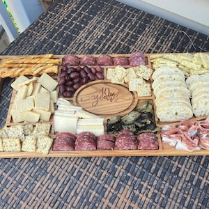 Puede incluir: Una tabla de servir de madera con una variedad de alimentos, que incluyen galletas, pan, queso, aceitunas, salami y prosciutto. La tabla está grabada con el texto "10 años".