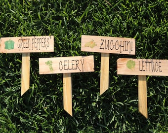 May include: Four wooden garden markers with painted illustrations of vegetables and the names "Green Peppers", "Zucchini", "Celery", and "Lettuce".
