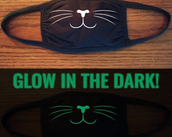May include: Black face mask with a white cat face design. The mask glows in the dark and the cat face design is green.