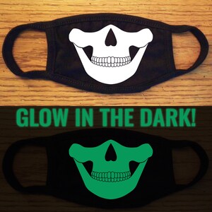 May include: Black face mask with a white glow-in-the-dark skull design. The skull has a large grin with teeth. The mask is shown in both regular and glow-in-the-dark conditions.