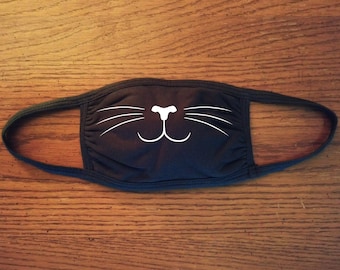May include: Black face mask with a white cat face design. The cat face has a nose, whiskers, and a smile.