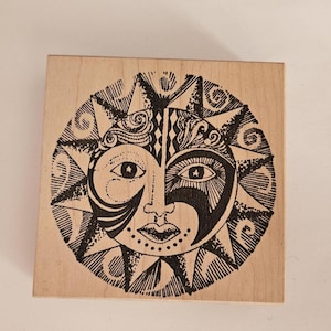 May include: A wooden stamp featuring a stylized sun face design. The sun's face is divided into two halves, one light and one dark, with intricate patterns and details. The stamp is square and made of light-colored wood.