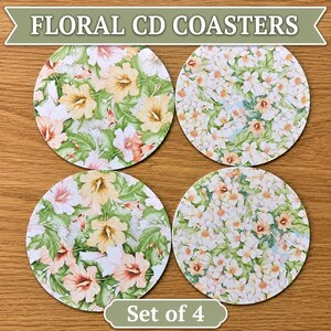 May include: A set of four circular coasters featuring floral patterns. Two coasters display large yellow and pink hibiscus-style flowers with green leaves, while the other two feature dense clusters of small white and peach blossoms with green foliage.