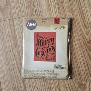May include: A packaged embossing folder by Sizzix with the text "Merry Christmas" in a decorative font. The folder is red and cream-colored, with the words "Texture Fades" and "Alterations" printed on the packaging. The product is designed for crafting.