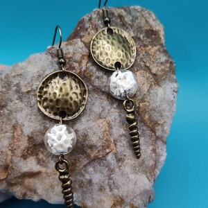 May include: A pair of dangle earrings with a hammered metal disc, a white bead, and a spiral-shaped pendant. The earrings are on a rock background.