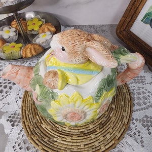 Puede incluir: Una tetera de cerámica con una figura de conejito, decorada con girasoles amarillos y hojas verdes. La tetera está sobre una estera tejida. En el fondo, una bandeja de dos niveles con pasteles.