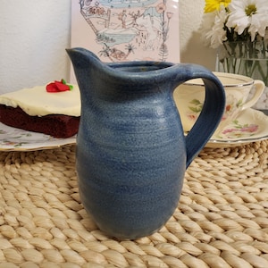 Puede incluir: Una jarra de cerámica azul con una superficie texturizada. La jarra tiene un cuerpo redondeado y un asa. Está sentada sobre un mantel tejido.