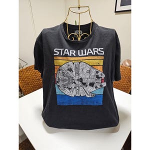 Può includere: Maglietta nera con una grafica Star Wars in stile vintage che presenta il Millennium Falcon su uno sfondo a strisce arcobaleno. Il testo "STAR WARS" è stampato sopra l'immagine.