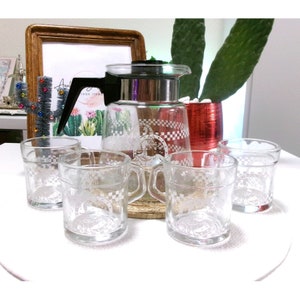 May include: A set of five clear glass cups and a glass pitcher with a checkered pattern and a silver lid. The cups and pitcher have a white design of a person riding a horse.