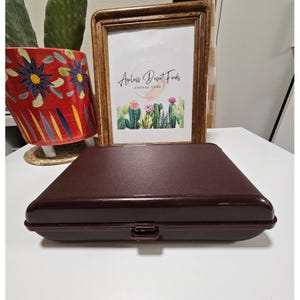 Puede incluir: Una caja rectangular de color marrón oscuro con cierre de pestillo. La caja está hecha de un material texturizado y tiene un borde ligeramente redondeado. La caja está sobre una superficie de color marrón claro.