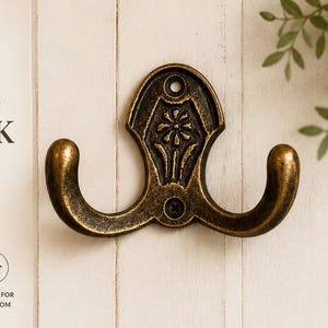 May include: A double wall hook with a dark bronze finish and a floral motif. The metal hook features two curved arms and a central plate with a stamped flower design. Text on the image reads: VINTAGE CHARM DOUBLE WALL HOOK, A timeless accent that brings warmth and character to any space, VINTAGE DESIGN, STURDY & DURABLE, EASY TO INSTALL, PERFECT FOR ANY ROOM, BEAUTIFUL, FUNCTIONAL, MADE TO LAST.