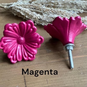 May include: Two magenta flower-shaped cabinet knobs with a silver screw. The knobs are on a wooden surface.