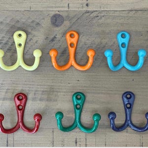 May include: A set of six double-pronged wall hooks arranged in two rows. The hooks are colored yellow, orange, light blue, red, green, and dark blue. Each hook features a vertical mounting plate with two screw holes and two curved arms.