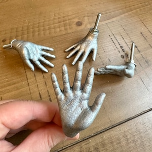 May include: Four silver hand-shaped drawer pulls with a brushed metal finish. The pulls are in various positions, with one being held in a hand.