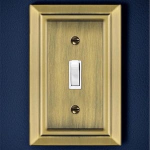 May include: A single light switch with a brushed brass finish and a white toggle switch. The switch is surrounded by a decorative brass frame.