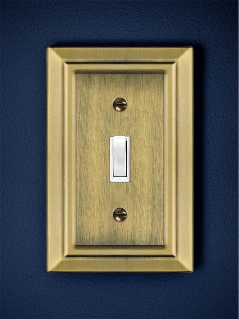 May include: A single light switch with a brushed brass finish and a white toggle switch. The switch is surrounded by a decorative brass frame.