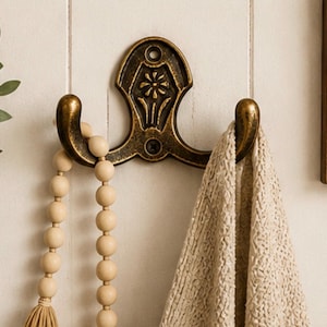 May include: A double-pronged wall hook with an antique brass finish and a central floral relief design. The hook features two curved arms, one holding a string of light-colored wooden beads and the other supporting a textured beige fabric cloth.