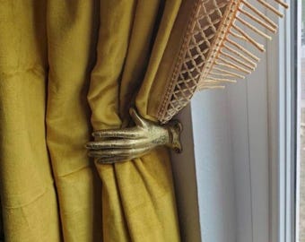 May include: A metallic gold-toned hand-shaped curtain tieback mounted to a wall, holding back a mustard yellow velvet curtain. The curtain features a decorative beige trim with a lattice pattern and long, dangling fringe along the top edge.