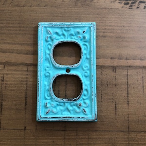 May include: A light blue, decorative outlet cover with an ornate design. The cover has a distressed finish, giving it a vintage look.