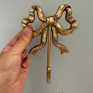 May include: A gold-colored metal wall hook featuring a decorative bow design at the top with a textured finish. The hook extends downward from the center of the bow. The text "Vintage Gold" is printed in black at the bottom of the image.