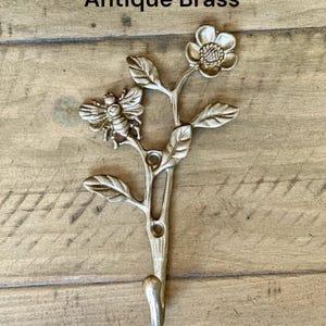 May include: An antique brass wall hook featuring a sculpted bee resting on a floral stem with leaves and a single flower at the top. The metal hook has two circular mounting holes in the center of the stem and the text Antique Brass above it.