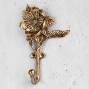 May include: A wall-mounted hook crafted from antique-toned brass, featuring a detailed three-dimensional flower bloom at the top with a single leaf extending to the right. The hook has two circular screw holes for mounting and a rounded tip.