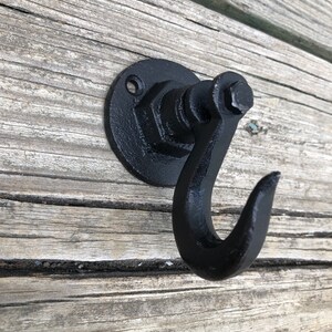 May include: Black metal hook with a round base and a single hook. The hook is attached to a wall with a screw.