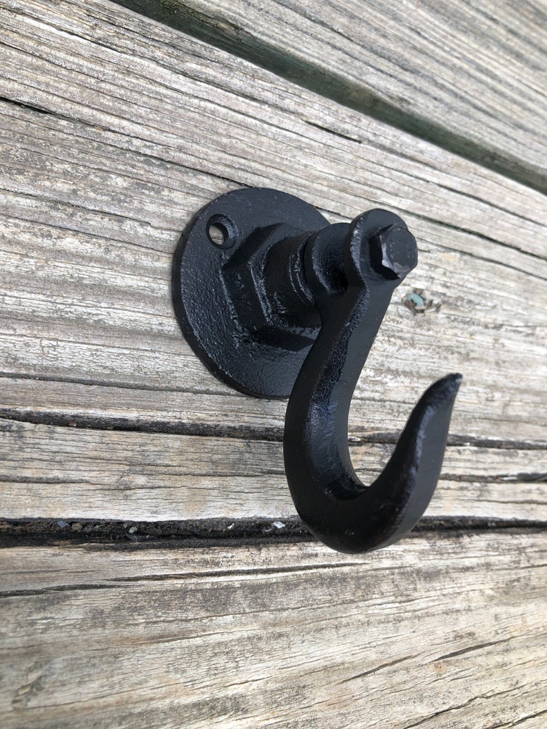 May include: Black metal hook with a round base and a single hook. The hook is attached to a wall with a screw.