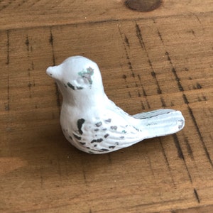 May include: A small white bird figurine with a distressed finish. The bird has a slightly curved beak and a rounded body with a textured surface.