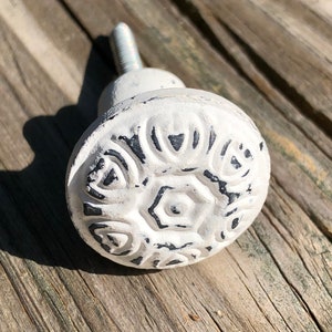 May include: White ceramic cabinet knob with a detailed floral design. The knob has a distressed finish and a silver screw.